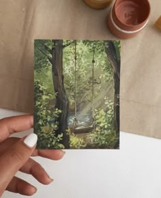 a person holding up a card with a swing in the woods next to some cups