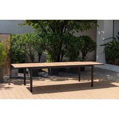 a wooden table sitting on top of a brick floor next to trees and bushes in front of a building