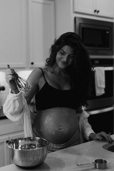 a pregnant woman is preparing food in the kitchen while holding a whisk and mixing bowl