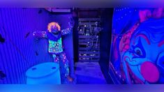 a clown standing next to a toilet in a room filled with blue and purple walls