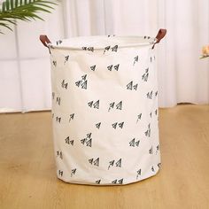 a white and black laundry basket with trees on it sitting on a wooden floor next to a potted plant