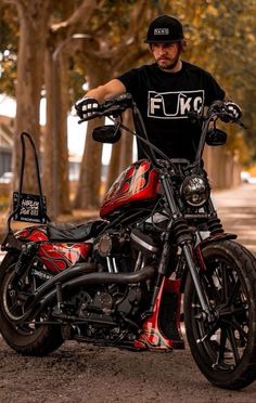 a man sitting on top of a red and black motor bike in the middle of a tree lined street