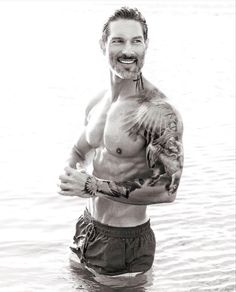 a man standing in the water with tattoos on his arm and chest, smiling at the camera