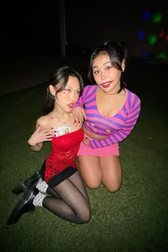 two young women sitting on the ground posing for a photo together at night with lights in the background