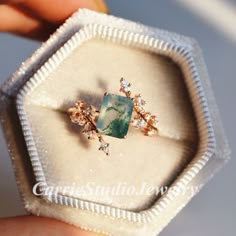 someone is holding an engagement ring in a box with some diamonds on it and a green stone surrounded by small white stones