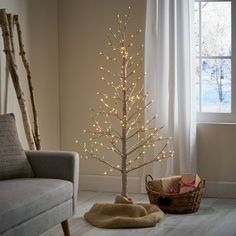 a small white christmas tree in a living room next to a chair and window with lights on it