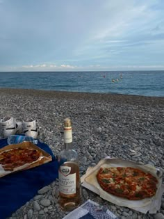 two pizzas sitting on top of a sandy beach next to the ocean with bottles of wine