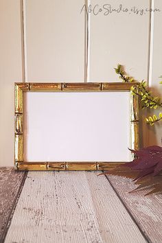 an empty gold frame sitting on top of a wooden table next to a vase with flowers