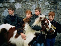 three boys are standing next to a small brown and white horse with long hair on it's back