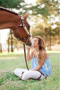 a woman sitting in the grass next to a brown horse and kissing her nose with it's bridle