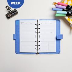 an open planner book next to some pens and pencils on a white table with a blue cover