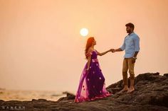 a man and woman holding hands on the rocks near the ocean at sunset or sunrise