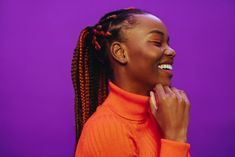 a woman with braids smiling and wearing an orange turtle - neck sweater against a purple background