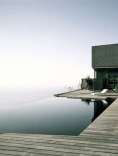 an empty swimming pool next to a wooden dock with chairs on the deck and water in the background