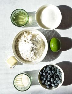 ingredients to make blueberry muffins laid out on a white tablecloth with shadows