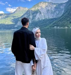 a man and woman standing on the edge of a body of water with mountains in the background