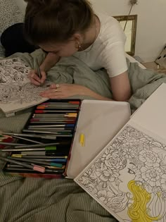 a woman laying in bed writing on a notebook with coloring books and pencils next to her