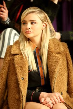 a woman sitting down in front of a crowd wearing a brown coat and black dress