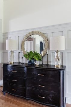 a black dresser with two lamps and a round mirror on the wall above it is decorated with greenery