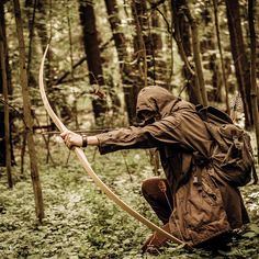 a man kneeling down in the woods with a bow and arrow on his back, aiming at something