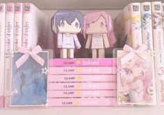 two anime dolls are standing on top of some books in a book shelf with pink covers