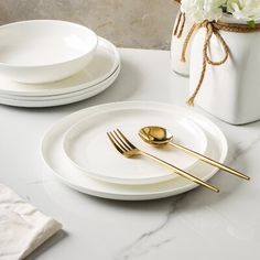 two white plates with gold forks and spoons sitting on a marble counter top next to a vase filled with flowers