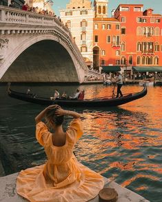 a woman in a yellow dress sitting on the edge of a river next to a bridge