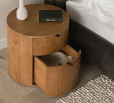 a round wooden table with two drawers and a white vase on the top, next to a bed