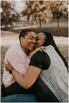 two women hugging each other while sitting on the ground in front of trees and grass