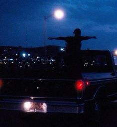 a man standing on the back of a pickup truck at night with his arms outstretched