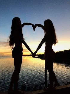 two girls making a heart shape with their hands by the water at sunset or dawn