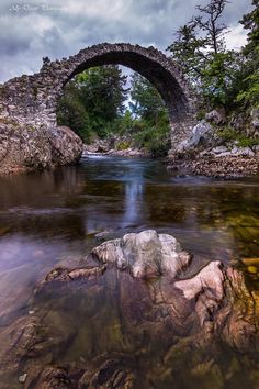 an old stone bridge over a river with rocks in the foreground and trees on either side