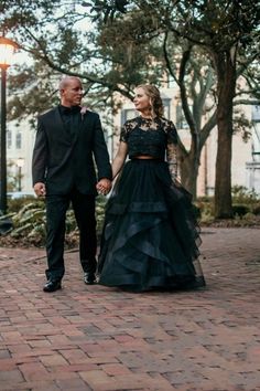 a man and woman holding hands walking down a brick walkway in front of some trees