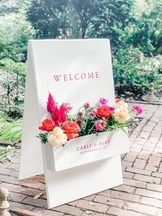 a welcome sign with flowers in it sitting on a brick walkway next to some bushes