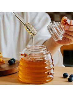 a person pouring honey into a jar with blueberries on the table next to it