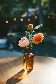 an orange and white flower in a brown vase on a wooden table with blurry lights behind it