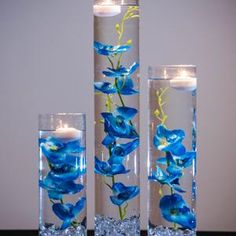 three tall glass vases with blue flowers and lit candles in them on a table