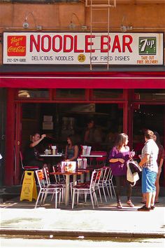 people are standing in front of a noodle bar with red awnings and tables outside