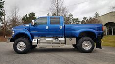 a large blue truck parked in a parking lot next to a building with trees on the other side