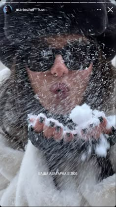 a woman wearing sunglasses and a black hat is blowing snow in the air with her hands