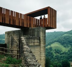 two people standing on top of a wooden bridge over a lush green valley with hills in the background