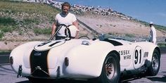 a man standing next to an old race car on the road with other people watching