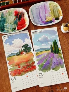 three calendars with paintings on them are sitting on a table next to two plates