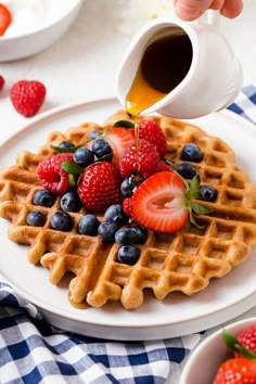 someone pouring syrup onto a waffle with strawberries and blueberries on the side