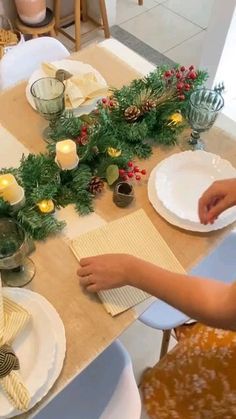 the table is set for christmas dinner with candles and napkins on it, along with evergreen garland