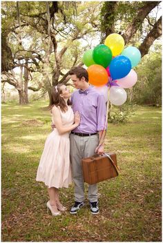 a man and woman kissing in the grass with balloons attached to their heads, holding a suitcase