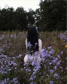 a woman sitting in the middle of a field with purple flowers on her knees and long black hair