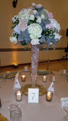 a tall vase filled with white and blue flowers on top of a table next to candles