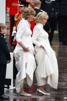 two women in white dresses and one man in uniform are walking down the street with other people