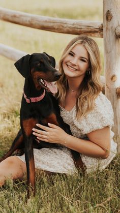 a woman sitting in the grass with her dog next to a wooden fence and looking at the camera